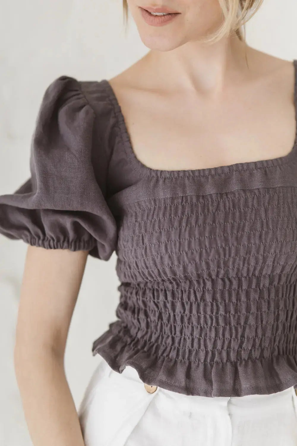 A close-up of a woman wearing a dark gray smocked puff-sleeve top with a square neckline, paired with white pants.