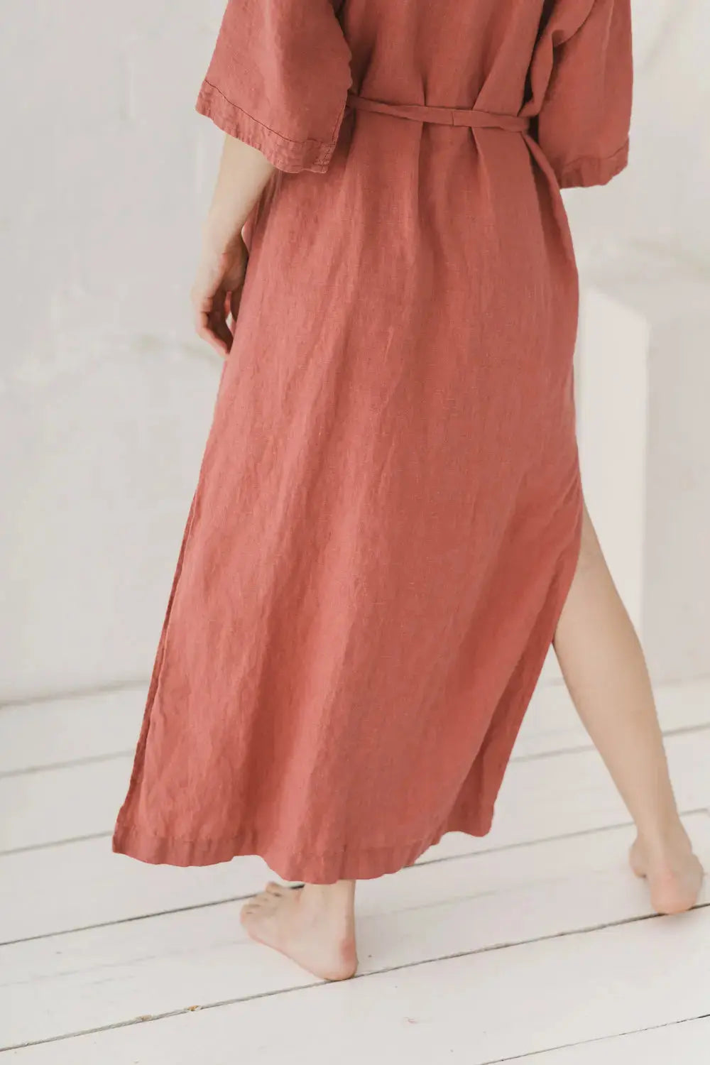 A woman wearing a dusty rose linen maxi dress with elbow-length sleeves and a matching tie belt, shown from the back as she walks barefoot on a white wooden floor.