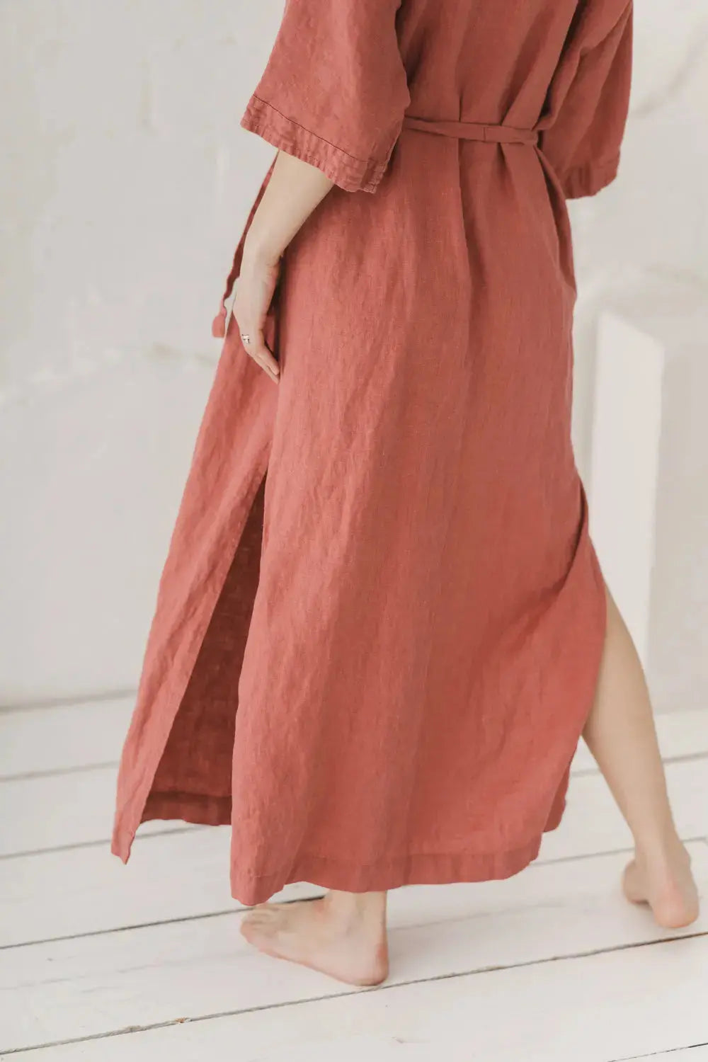 A person wearing a dusty rose linen maxi dress with a tie waist belt and side slits, shown from the back in a bright, minimalist room with white wooden floors.