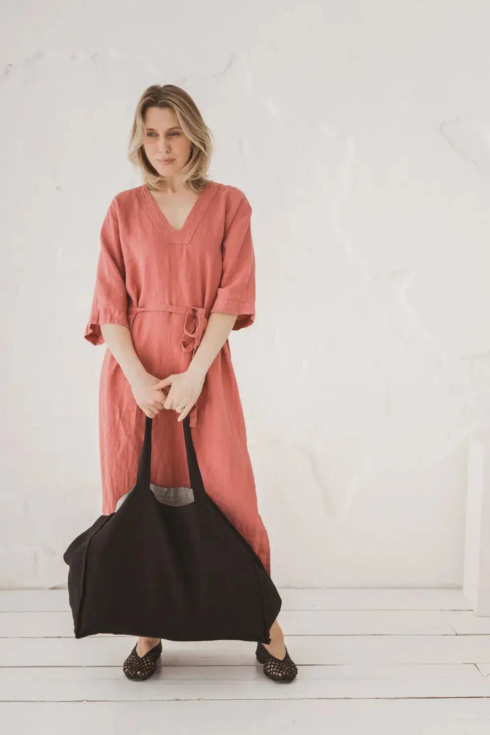 A woman wearing a dusty rose linen dress and holding a large black canvas tote bag, standing against a white textured wall.