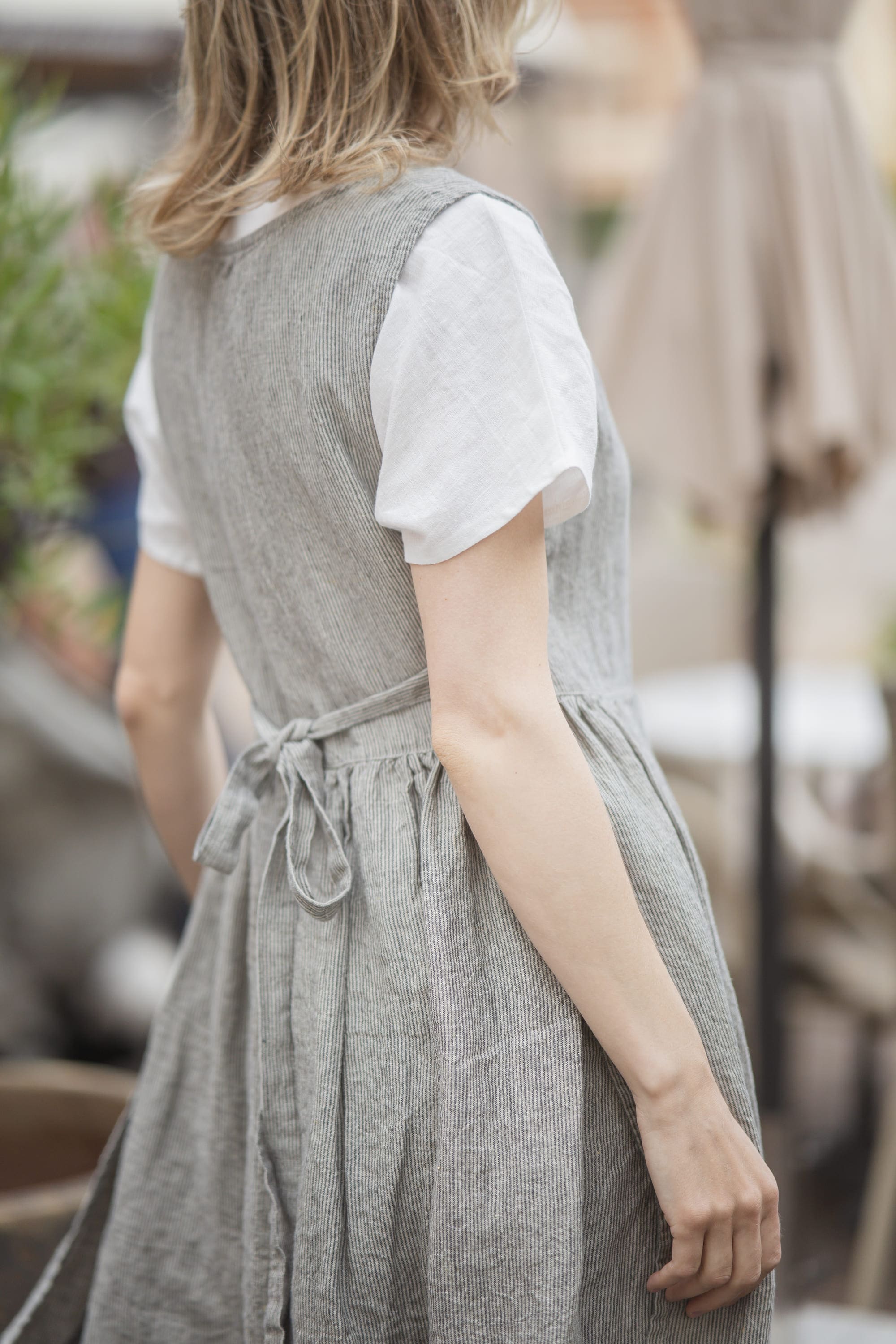A woman shown from behind wearing a light gray linen pinafore dress over a white short-sleeve t-shirt, with a matching gray tie belt at the waist.