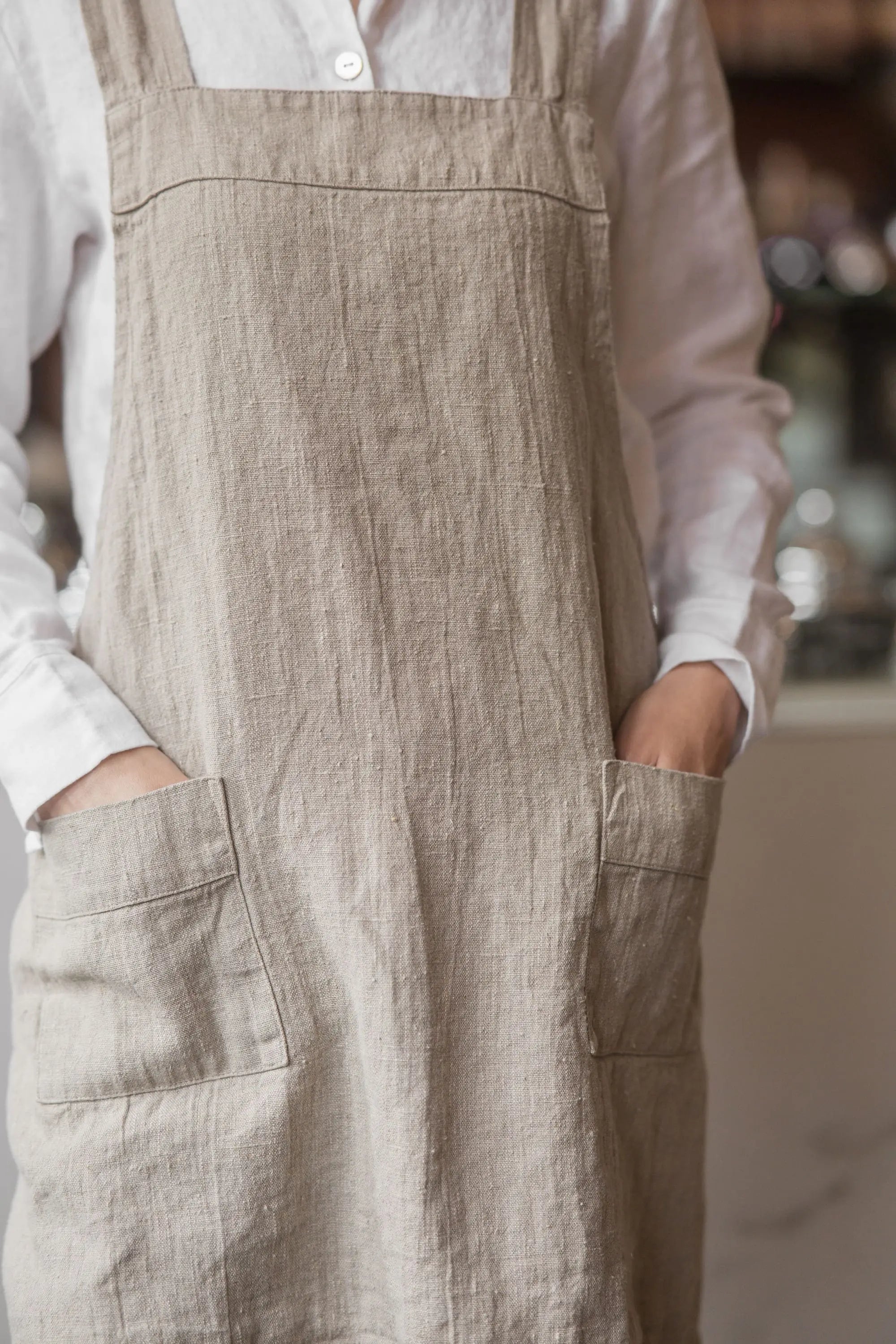 Close-up of two patch pockets on a natural linen apron, with hands resting inside each pocket, showing the linen's woven texture.