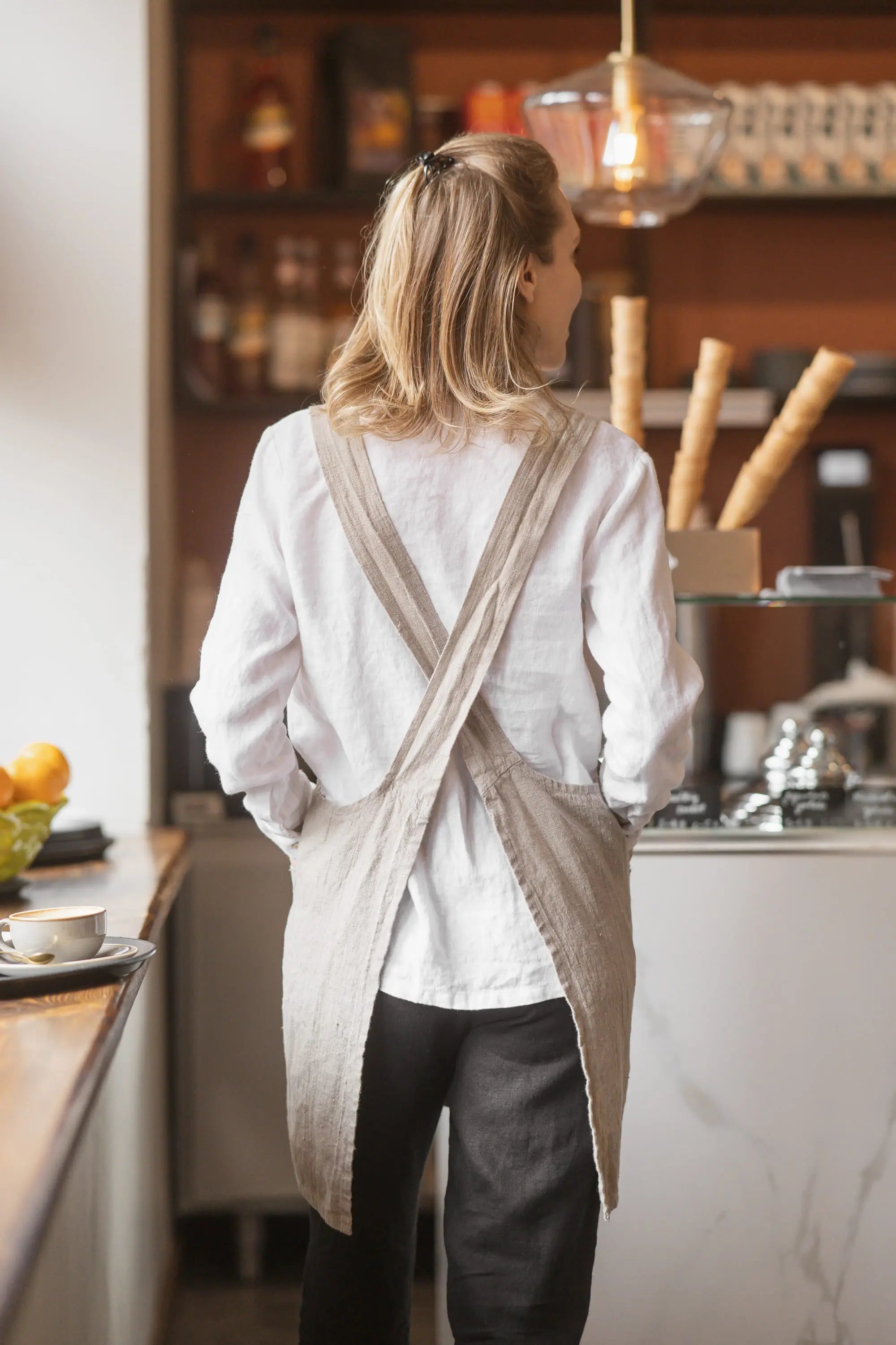 Back view of a natural linen apron showing the straps crossed in an X shape between the shoulders, worn over a white shirt.