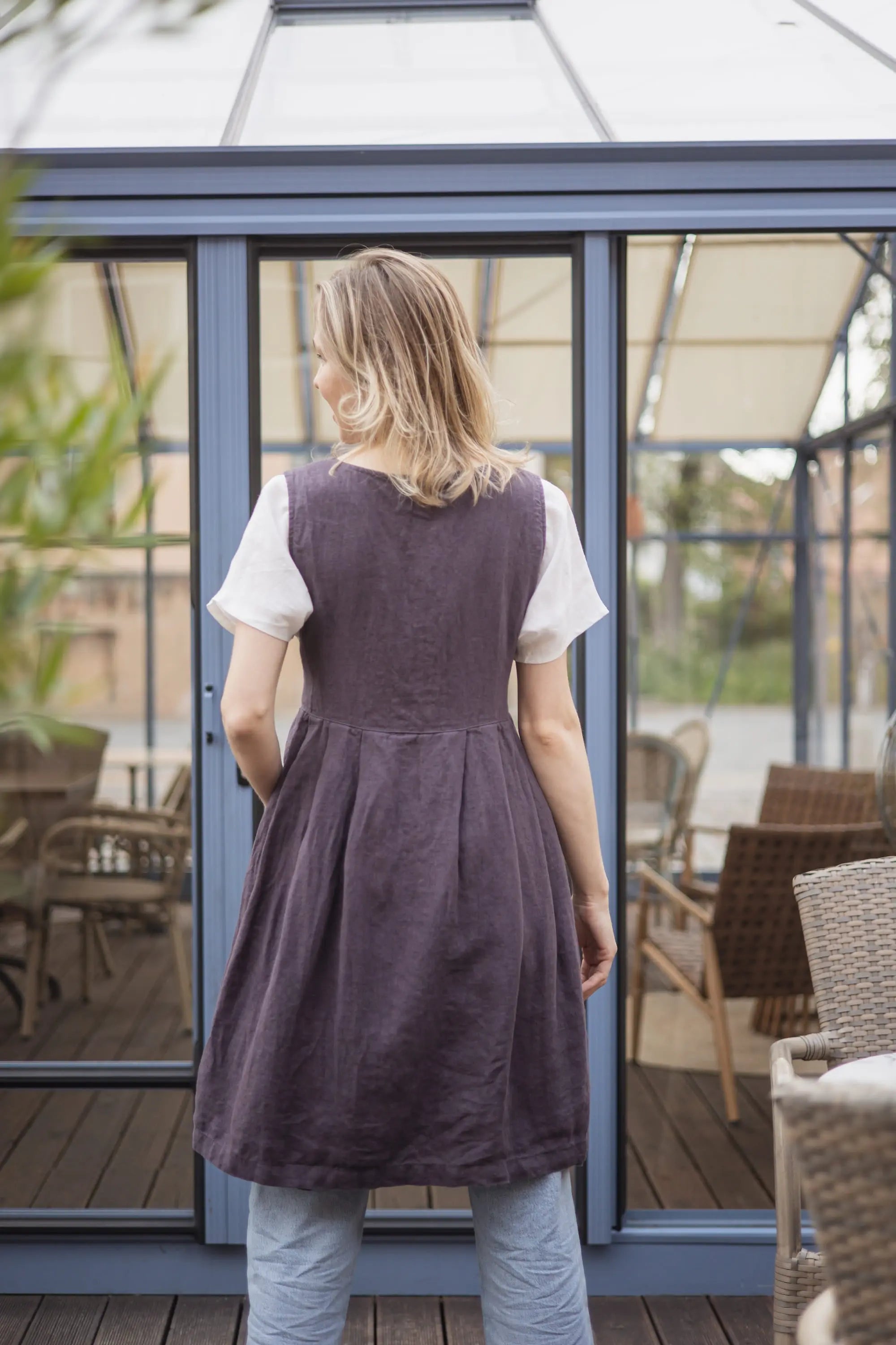 Woman viewed from behind wearing a plum linen pinafore apron with a gathered pleated skirt, worn over a white t-shirt and jeans in front of glass sunroom doors.