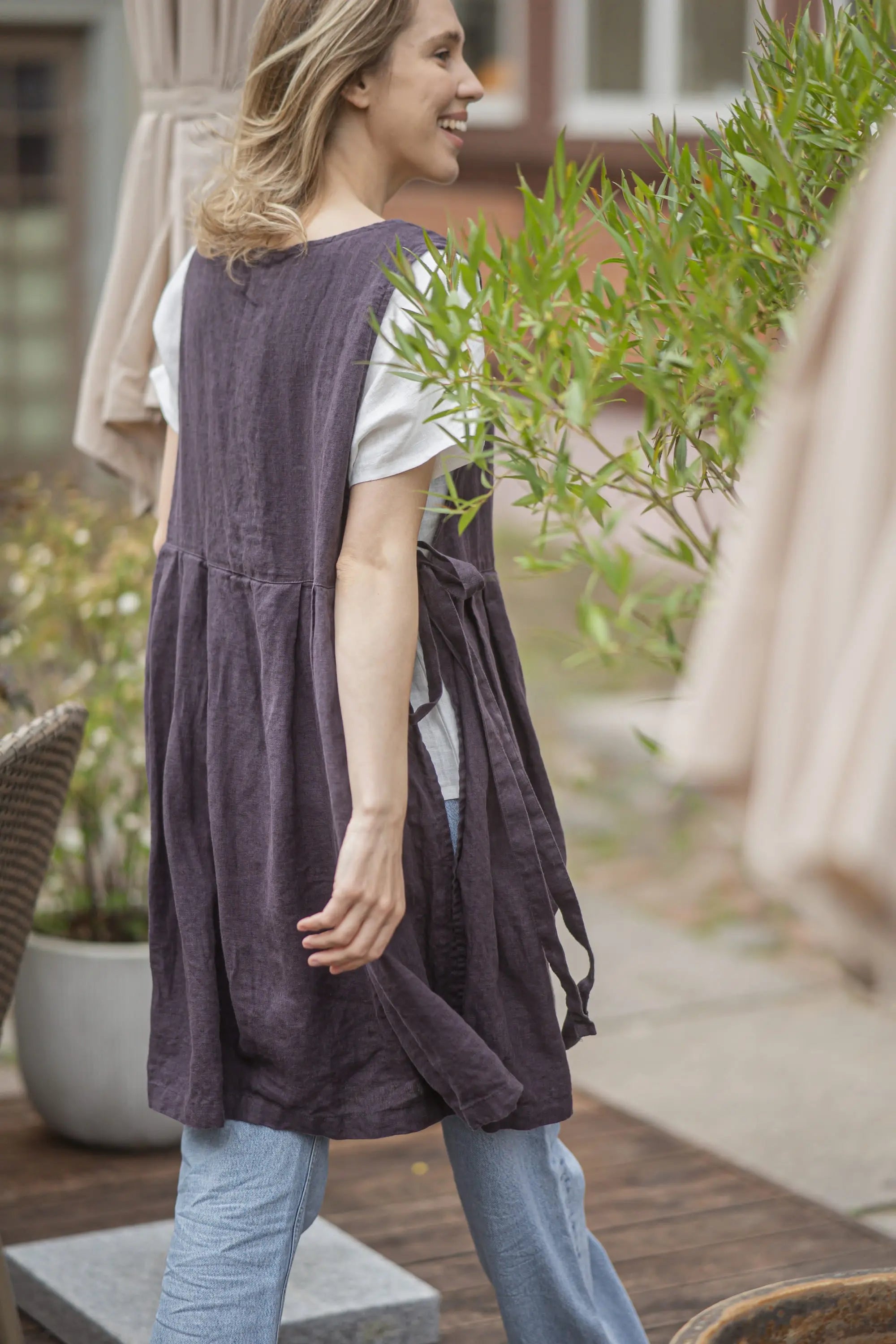 Woman walking outdoors past a bamboo plant, wearing a plum linen pinafore apron with loose tie straps at the side and a gathered pleated skirt.