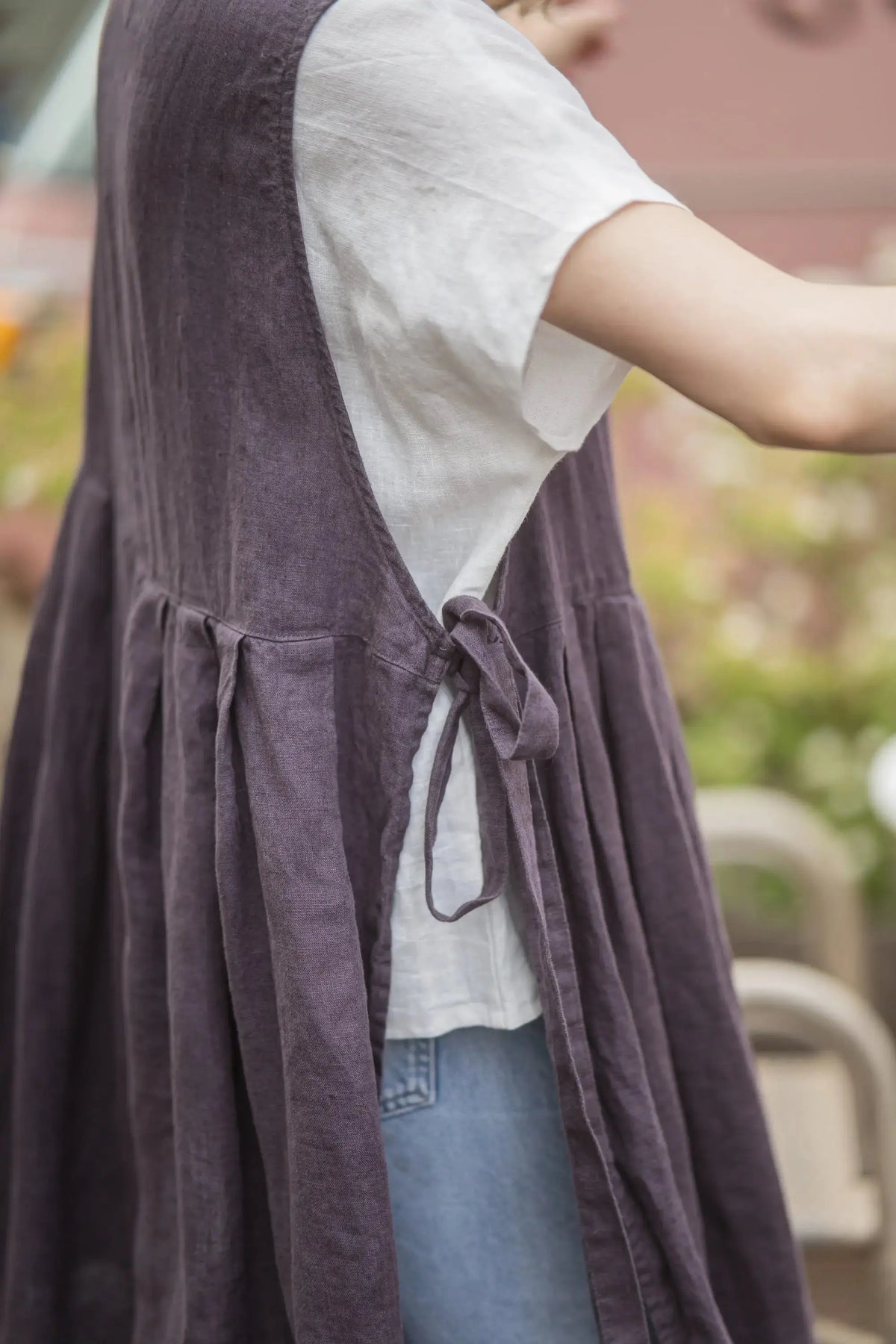 Close-up of the side tie bow and gathered pleats on a plum linen pinafore apron, worn over a white t-shirt with denim jeans visible underneath.