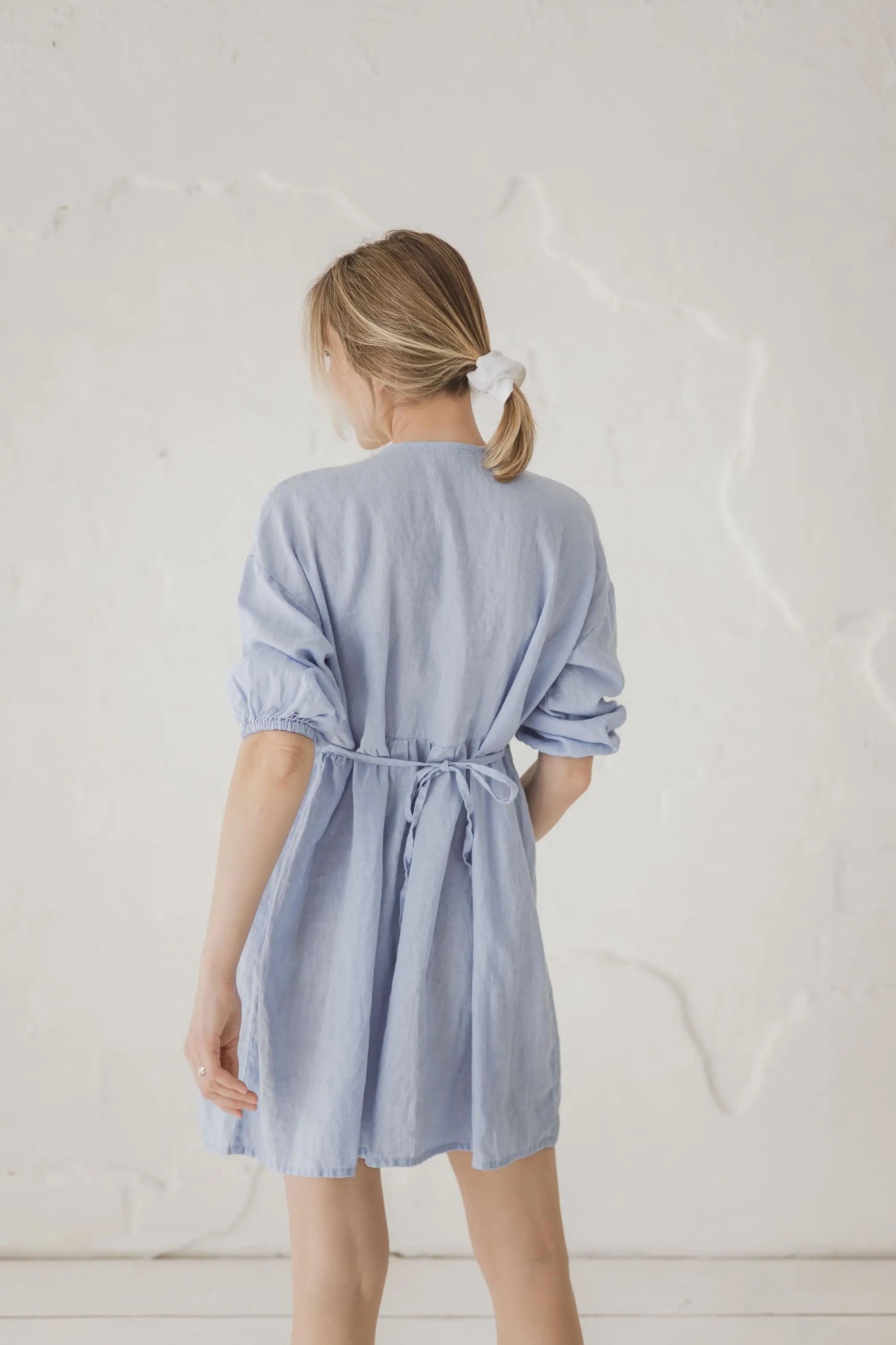 A woman viewed from behind wearing a light blue linen shirt dress with a tie waist, standing against a textured off-white wall.