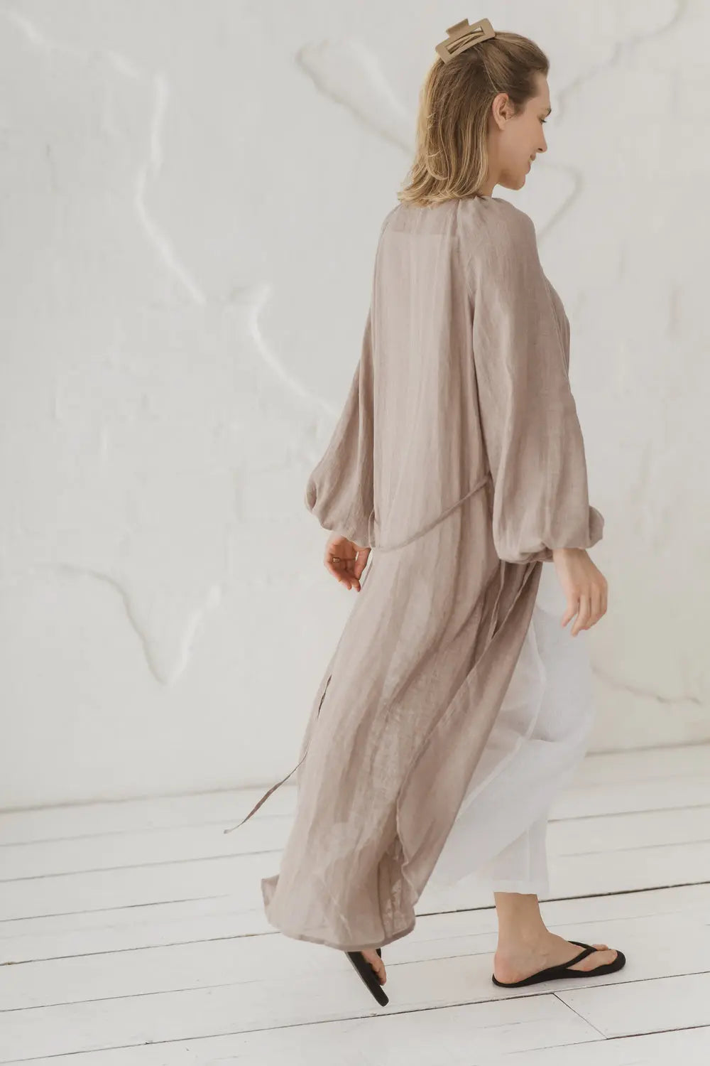 Side and back view of a woman walking in a natural linen kaftan with waist tie, styled with white linen trousers