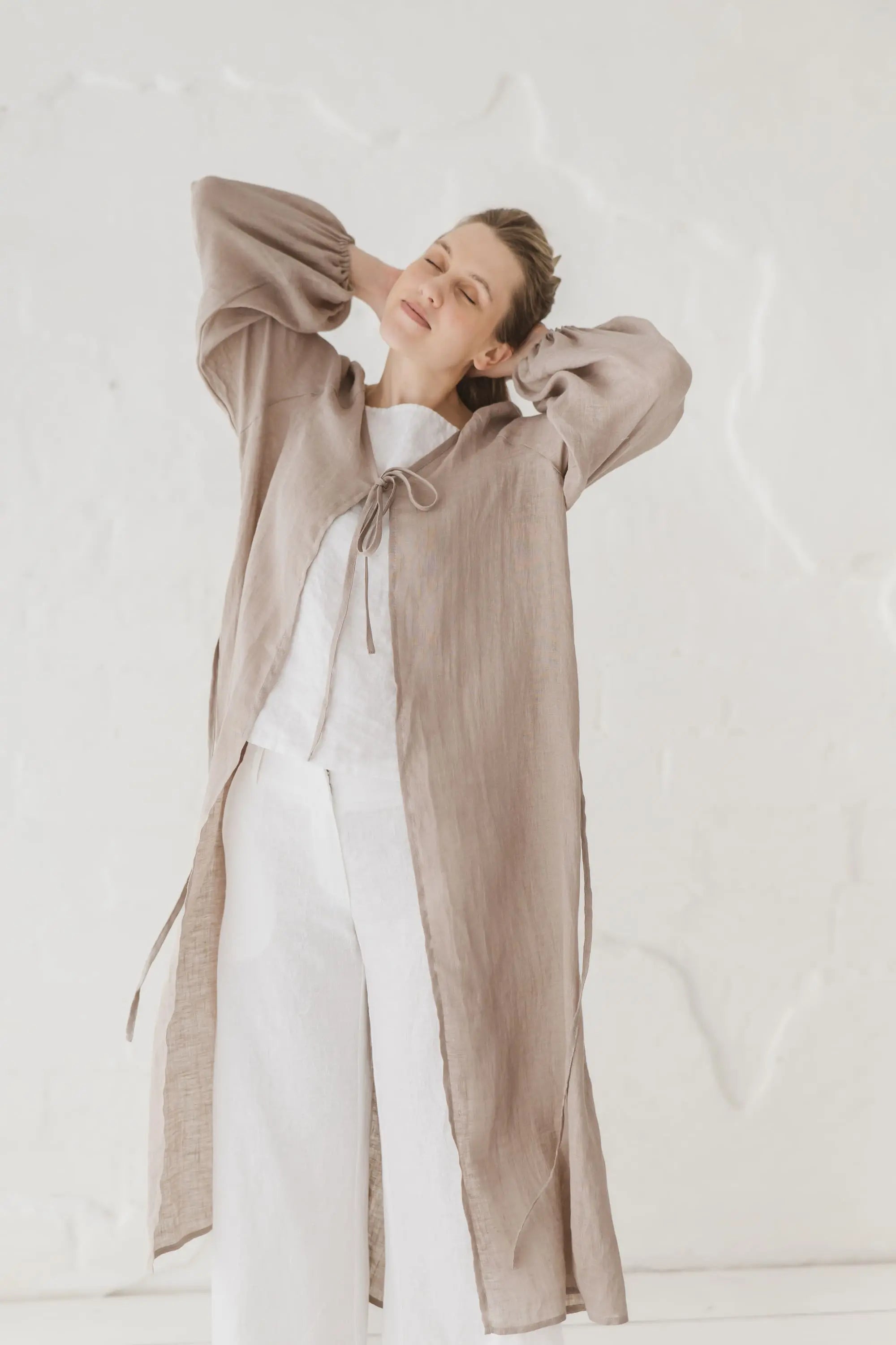 Woman in a natural linen kaftan with tie front and waist tie, arms raised, styled with white linen trousers