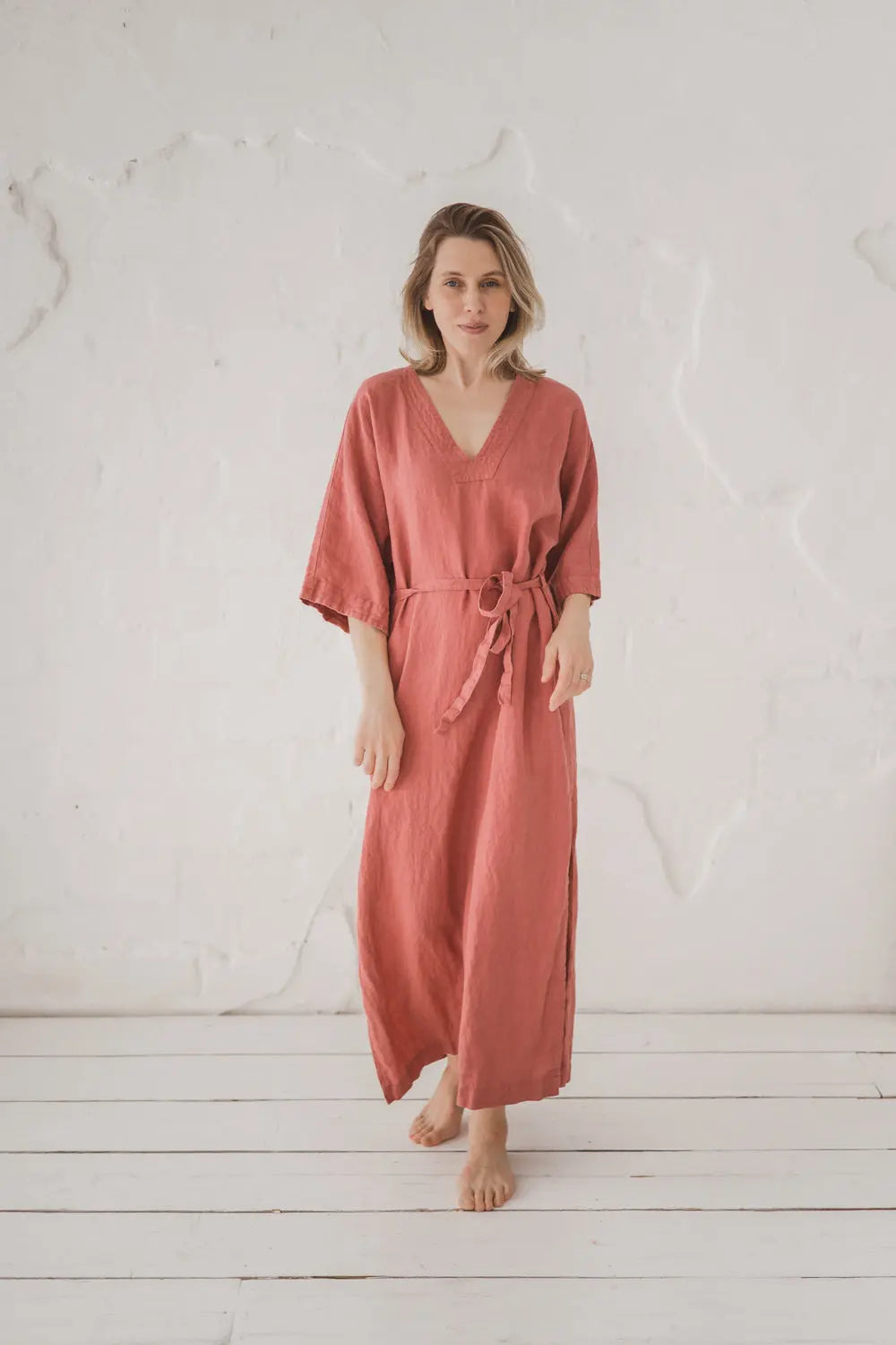A woman wearing a dusty rose linen kaftan dress with a tie waist belt, standing barefoot on a white wooden floor against a textured white wall.