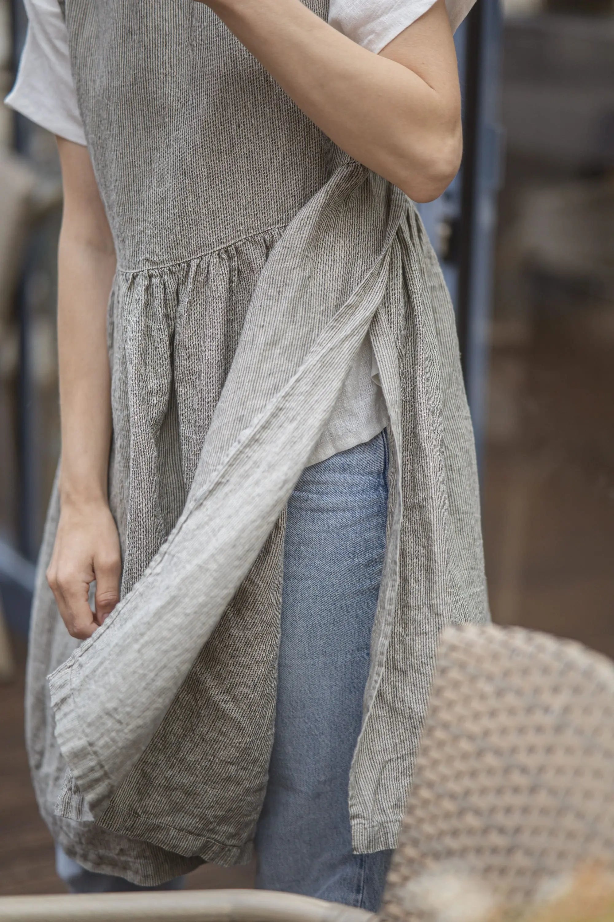 A person wearing a light gray linen apron over a white t-shirt and blue jeans, holding the apron's ties to adjust it.