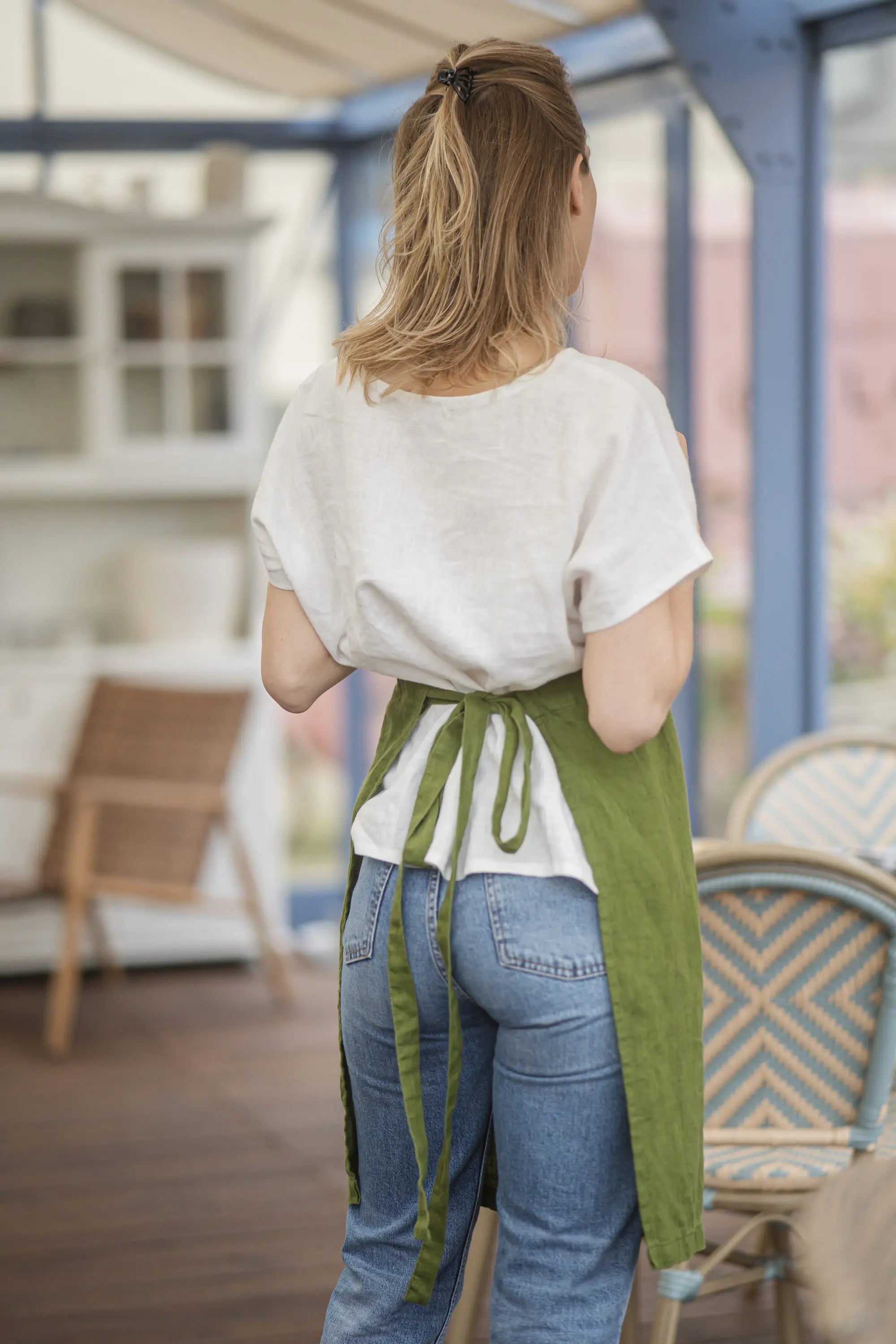 Back view of the olive green linen apron tied in a bow at the waist, worn over a white linen top and blue jeans
