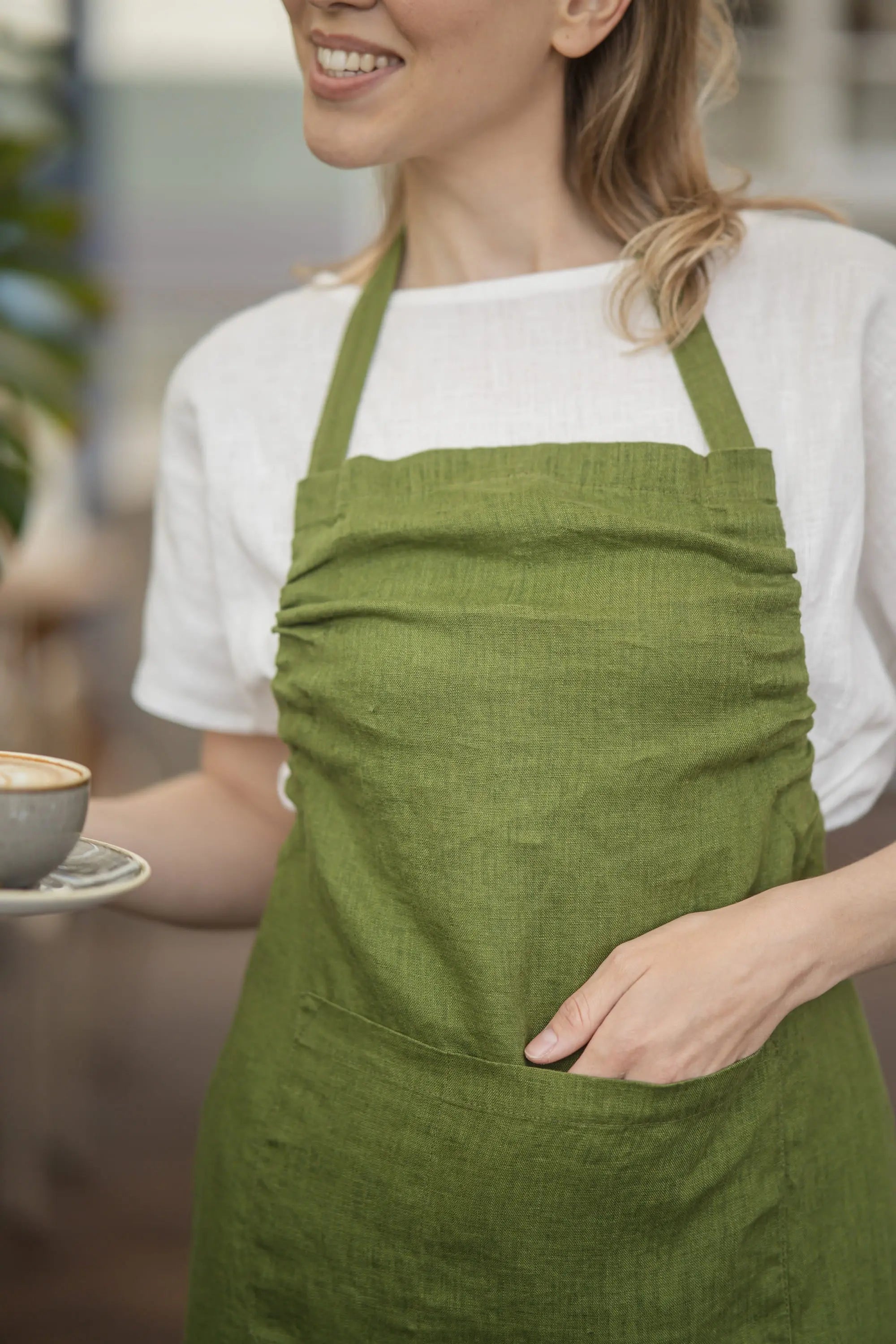 Close-up of the gathered bib and front patch pocket on the olive green linen apron, with a hand resting inside the pocket and linen fabric texture visible