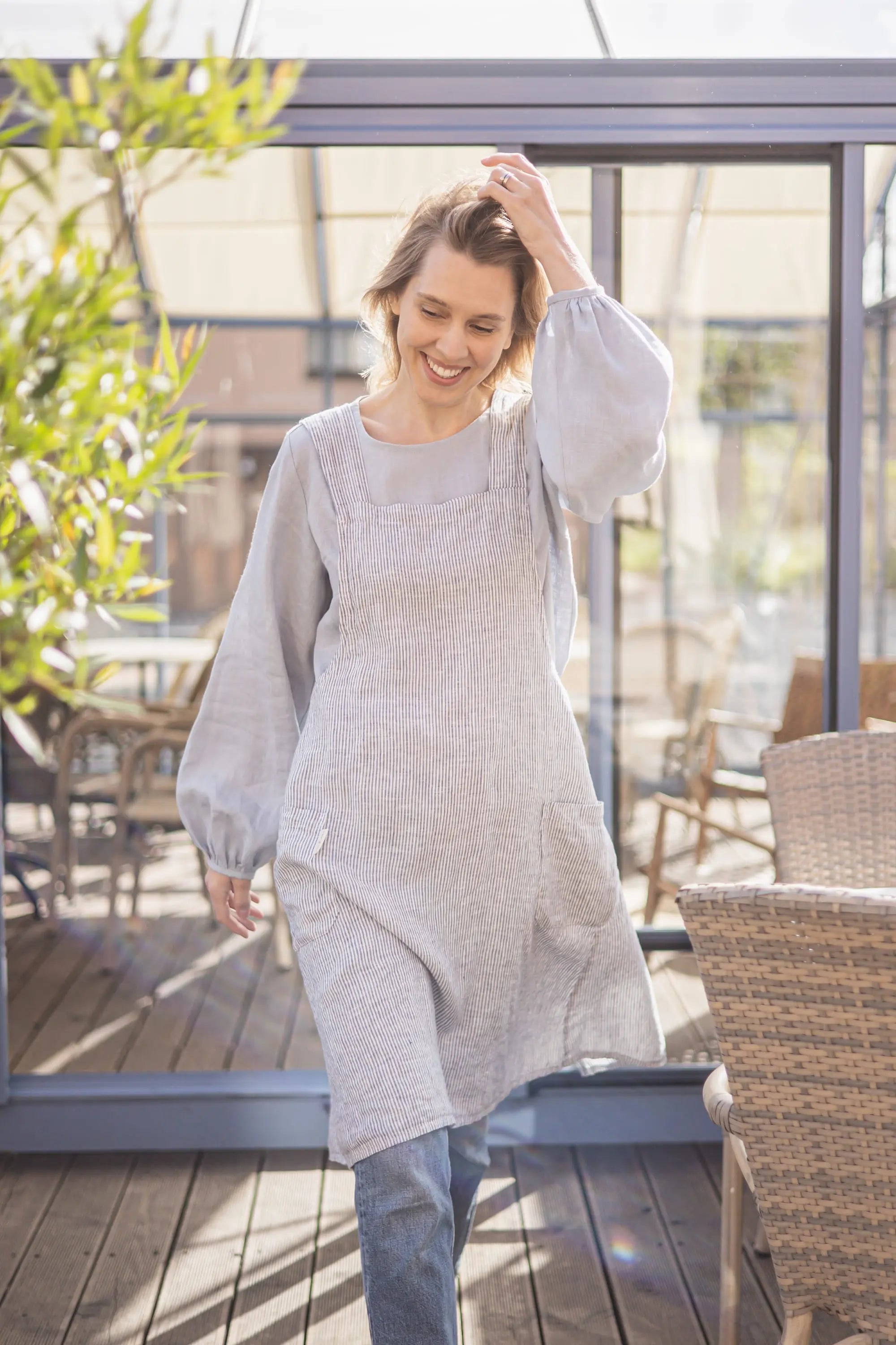 Woman walking on a sunlit terrace wearing a striped linen crossback apron with a visible front pockets, smiling with one hand touching her hair
