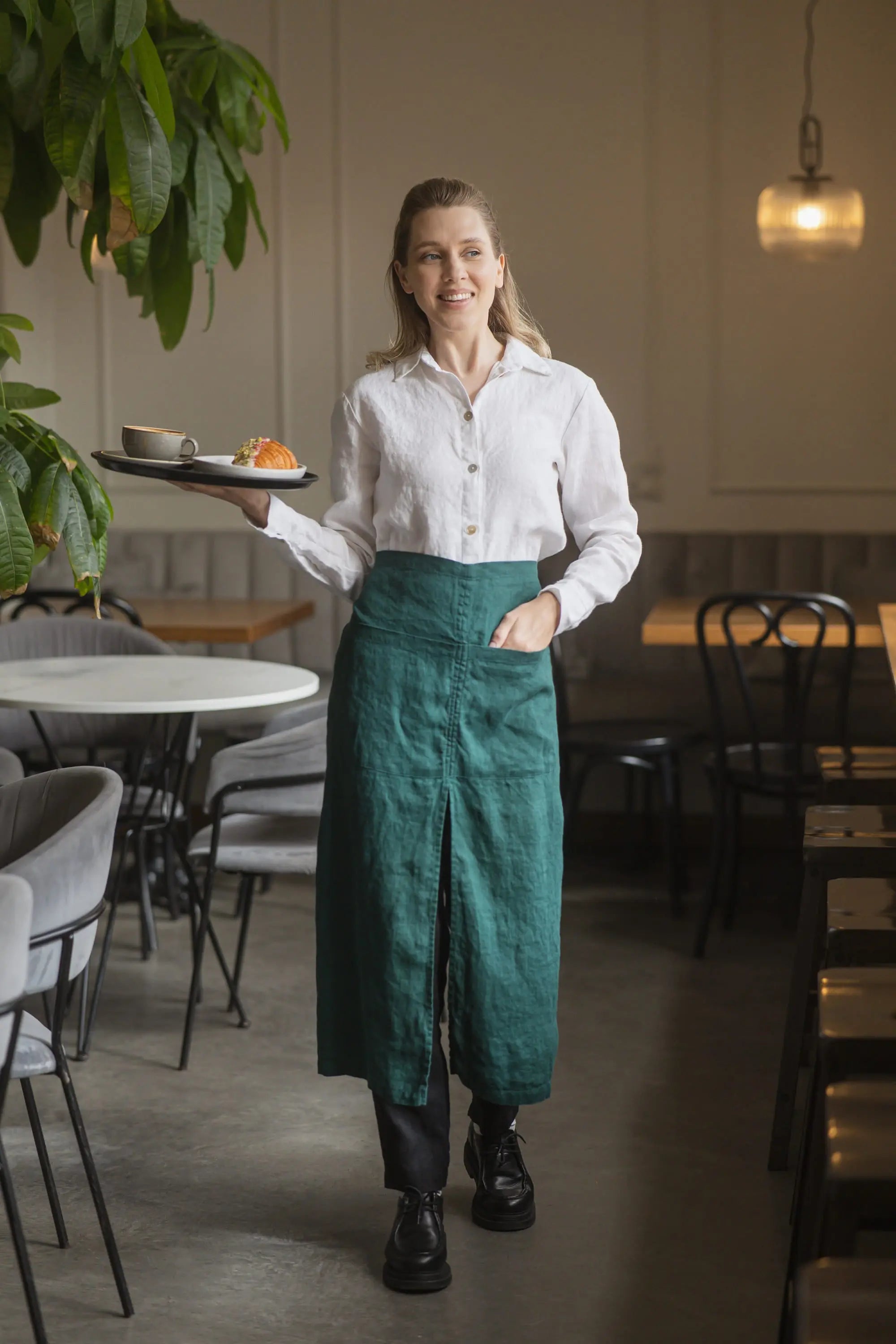 Woman standing in a café wearing a dark green linen waist apron over a white button-down shirt, holding a serving tray with a coffee cup and pastry.