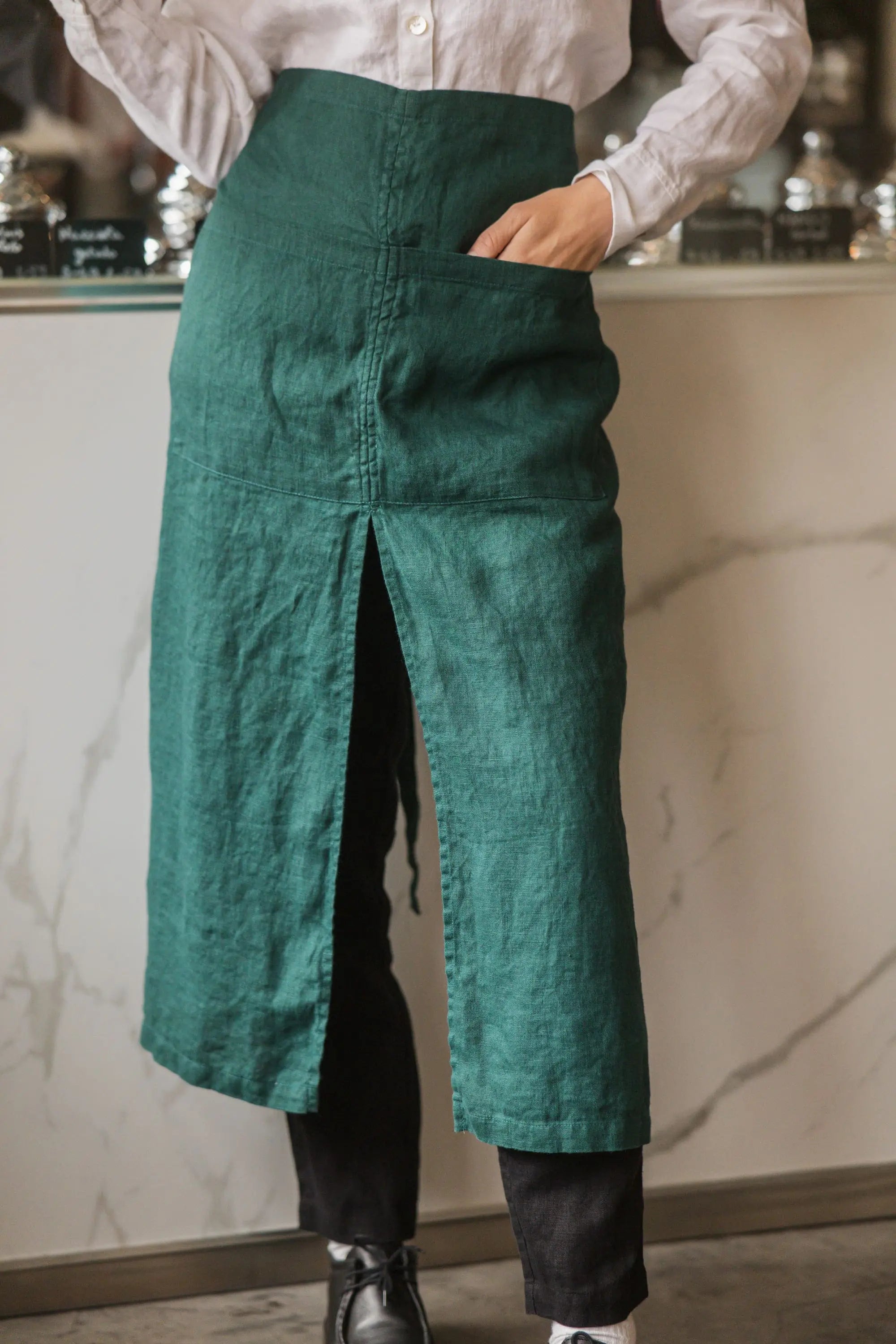 Close-up of the front slit and hip pocket on a dark green linen waist apron, hand tucked inside, with a gelato counter blurred in the background.