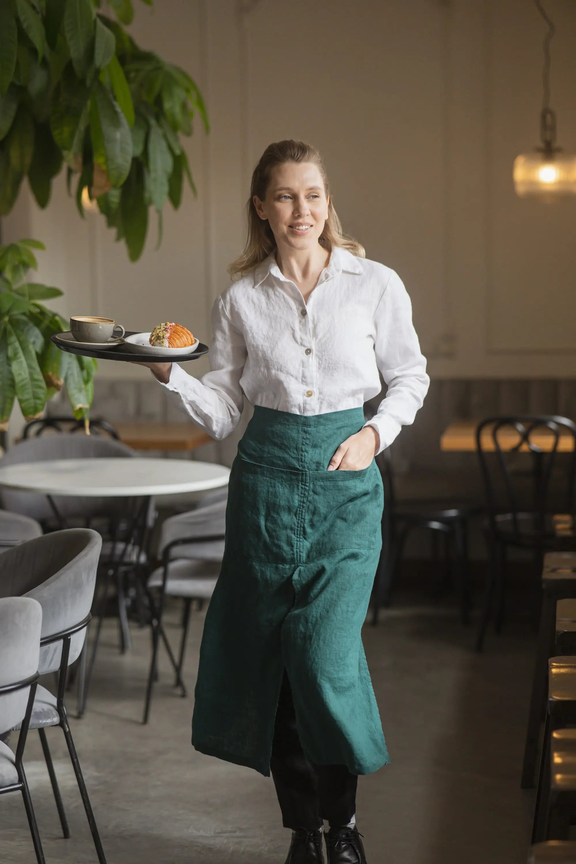 Close-up of a dark green linen waist apron showing the high waistband and hip pocket, with a hand tucked inside while holding a serving tray against a café backdrop.