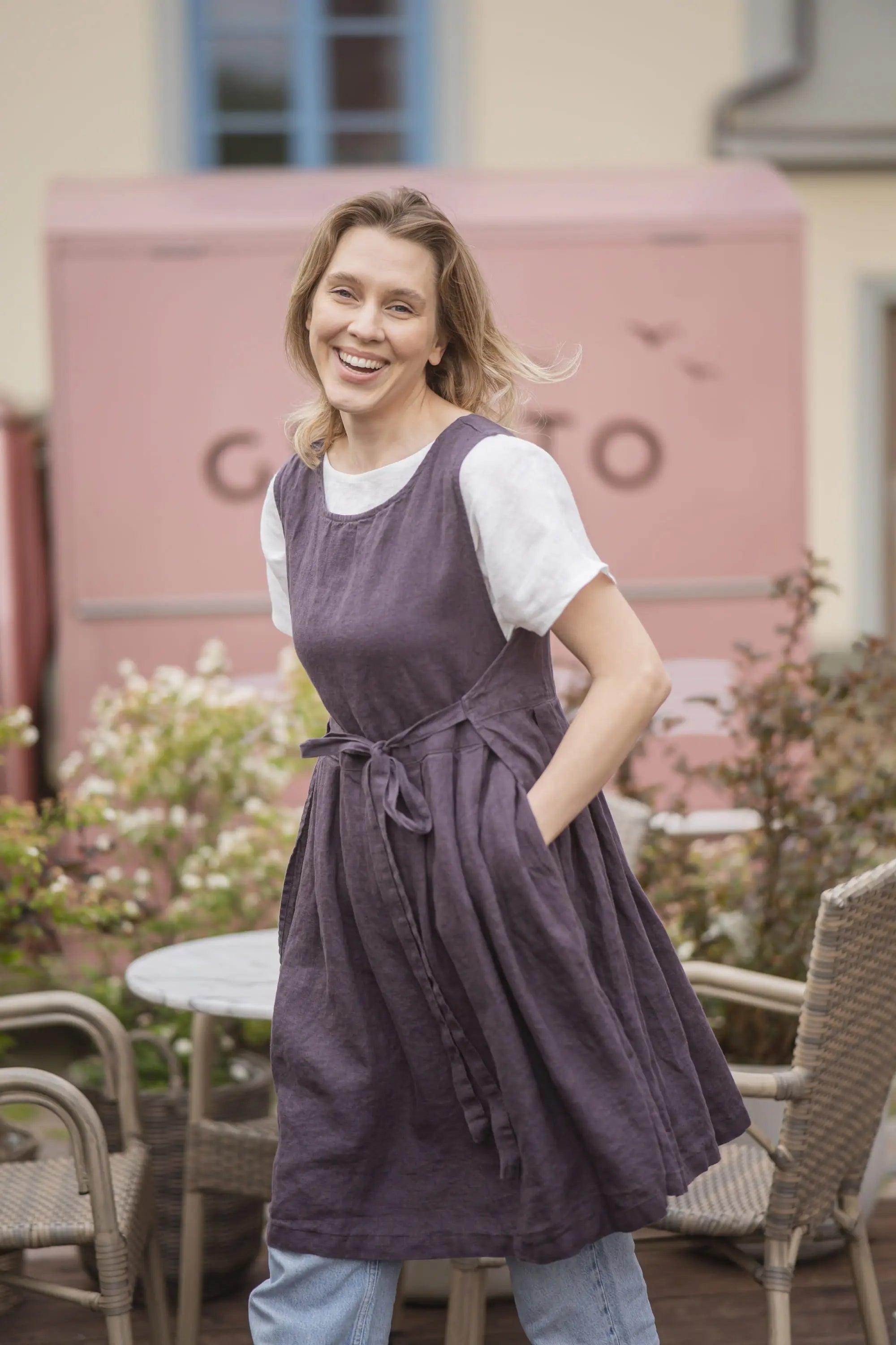 Woman wearing a plum linen pinafore apron with two side pockets, worn over a white t-shirt and light denim jeans.