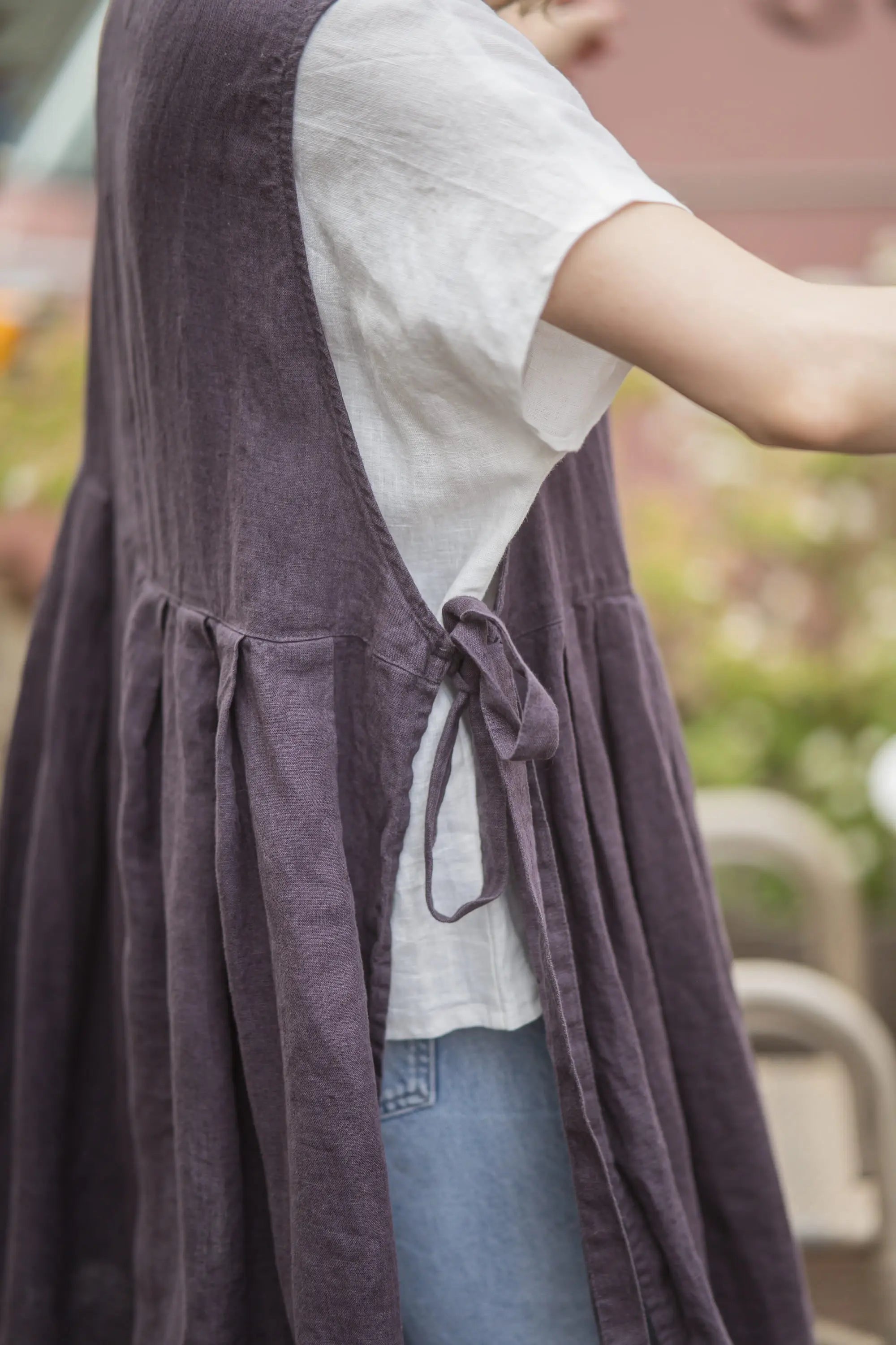 Close-up of the side tie bow and gathered pleats on a plum linen pinafore apron, worn over a white t-shirt with denim jeans visible underneath.
