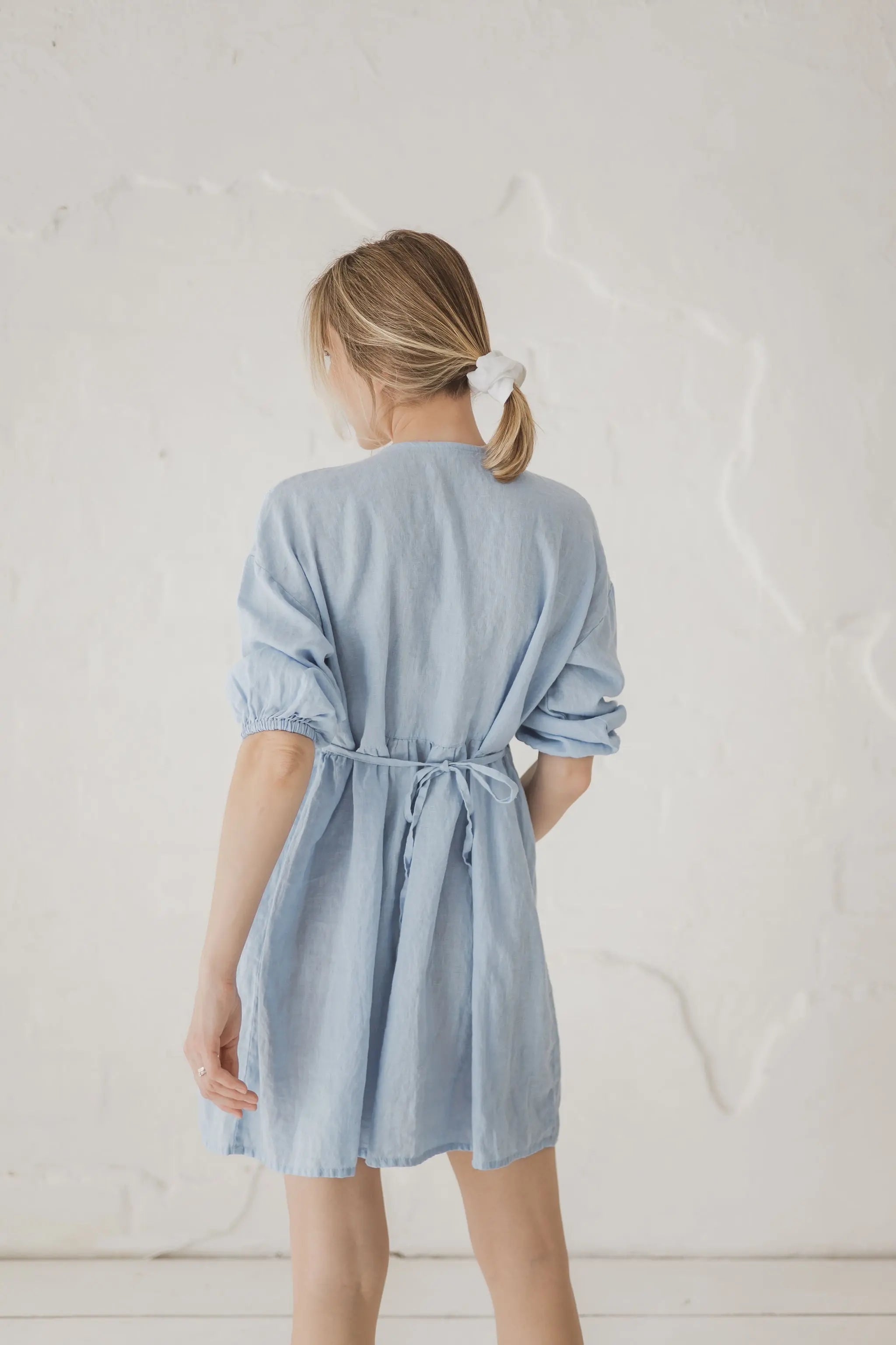 A woman viewed from behind wearing a light blue linen shirt dress with a tie waist, standing against a textured off-white wall.