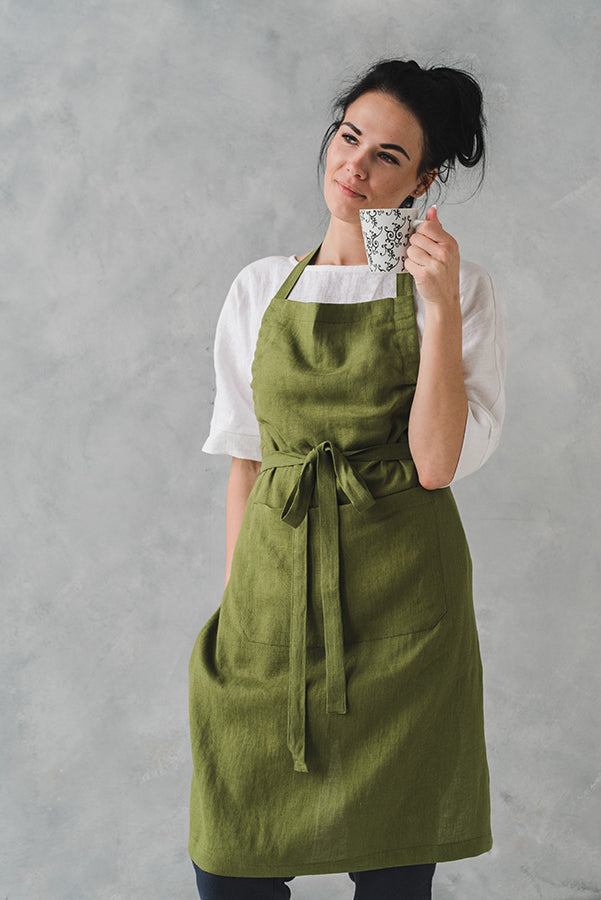 woman holding a cup of tea while wearing a beautiful green moss linen apron and a white linen blouse underneath