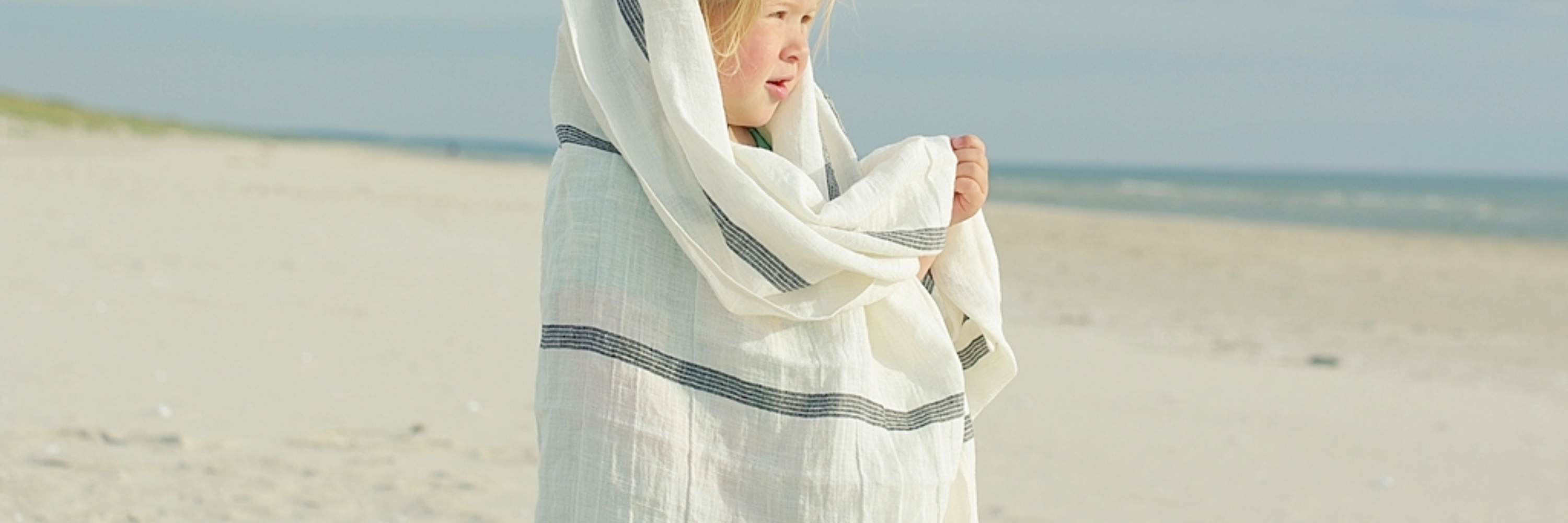 A girl standing on a beach wrapped in a beautiful linen huckaback towel.