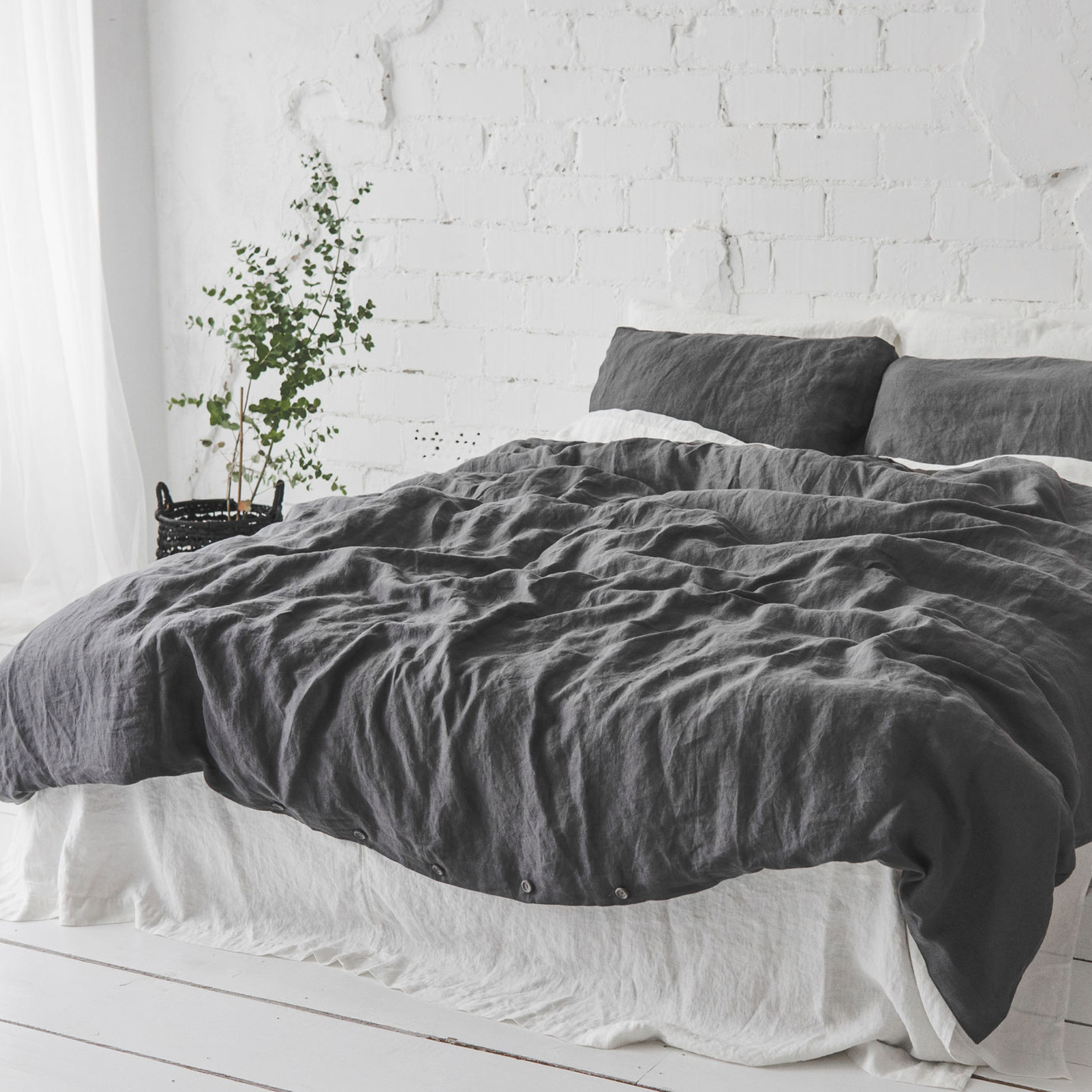 A made up bed with a dark gray liinen cover with a white flat sheet in a white room.