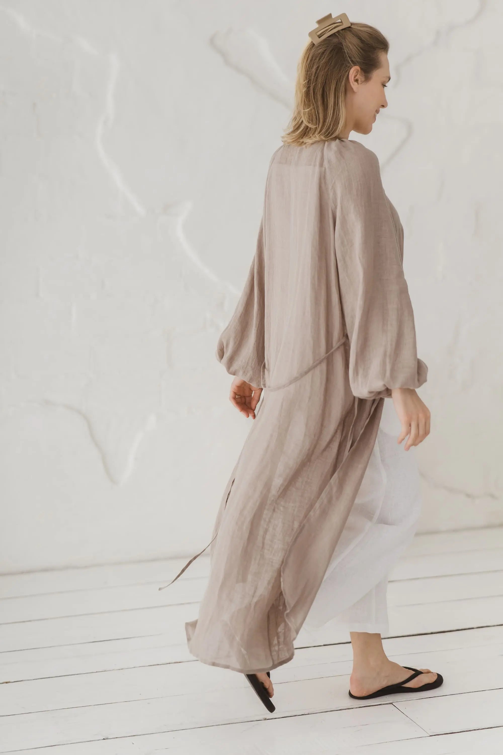 Side and back view of a woman walking in a natural linen kaftan with waist tie, styled with white linen trousers