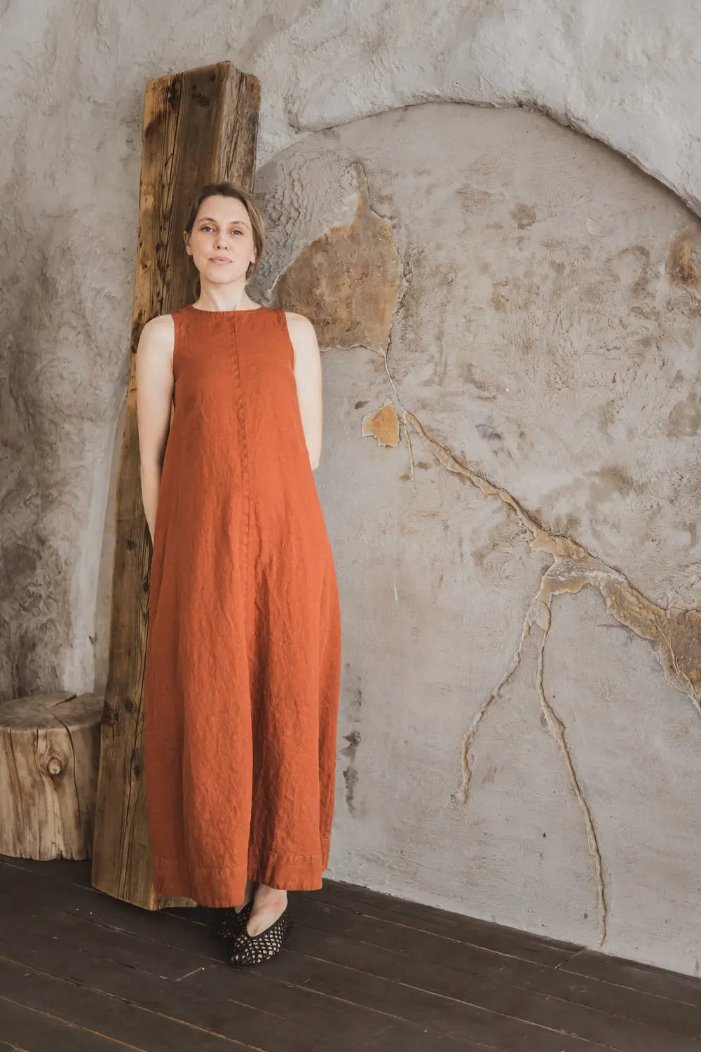 Woman in a sleeveless rust linen maxi dress with A-line silhouette leaning against a wooden beam, full length front view