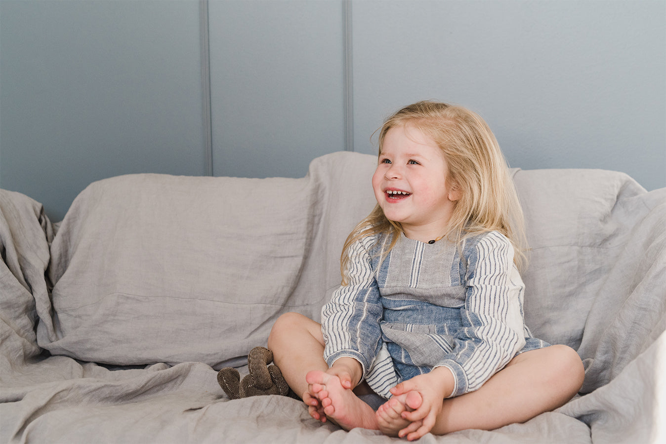 a toddler having fun sitting on a sofa with a linen cover and she is wearing a linen dress in blue stripes